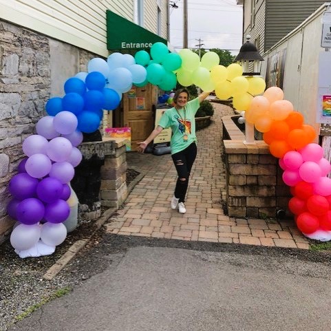 A welcoming person under a big pride balloon archway.